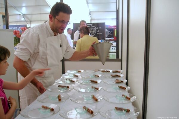 Chef in white pours from a metal funnel into small glass bowls arranged on a long table at a live cooking demo, a child watching nearby.