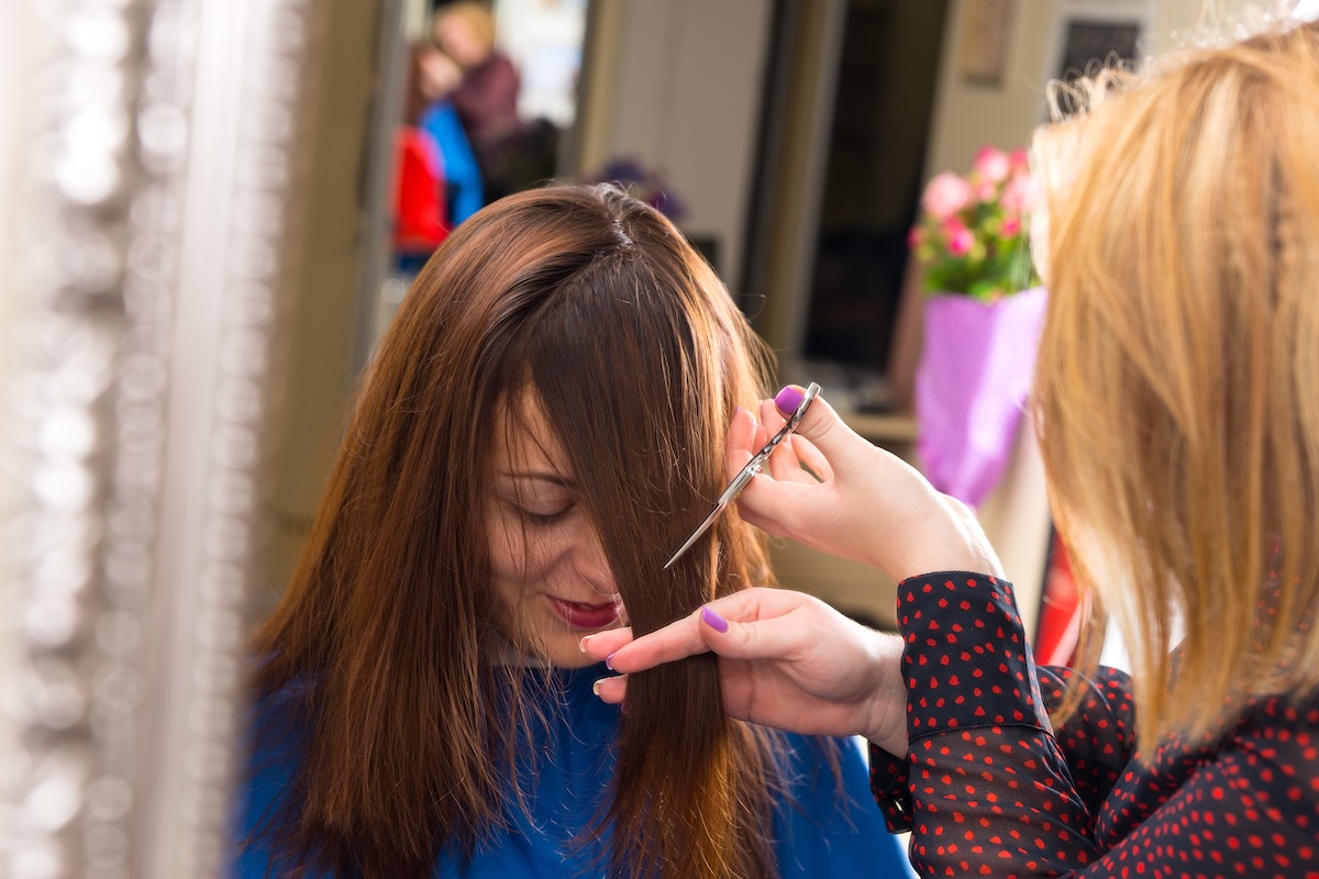 Close Up of Blond Stylist Cutting Hair of Young Brunette Client - Hair Dresser Cutting Angled Bangs into Hair of Young Female Client in Salon