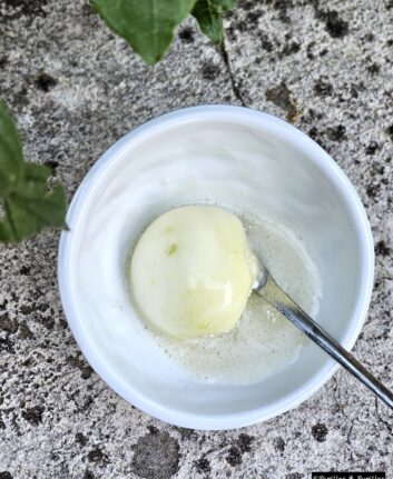 Peeled potato soaking in soapy water inside a white bowl on a rocky outdoor surface with a silver spoon nearby.