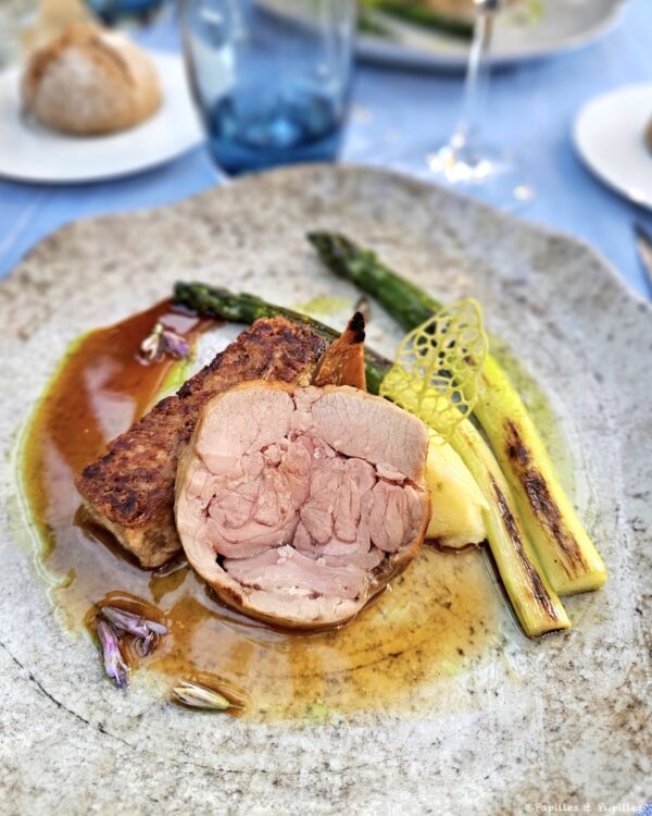 Sliced roasted pork with crackling, accompanied by charred leeks, yellow purée, and a delicate lattice crisp on a gray plate.