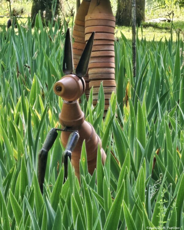 Copper rabbit sculpture with long ears standing among tall green garden leaves.