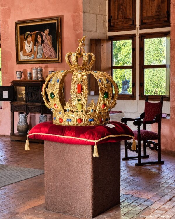 Gold royal crown encrusted with colorful jewels resting on a red velvet cushion atop a pedestal in a museum room