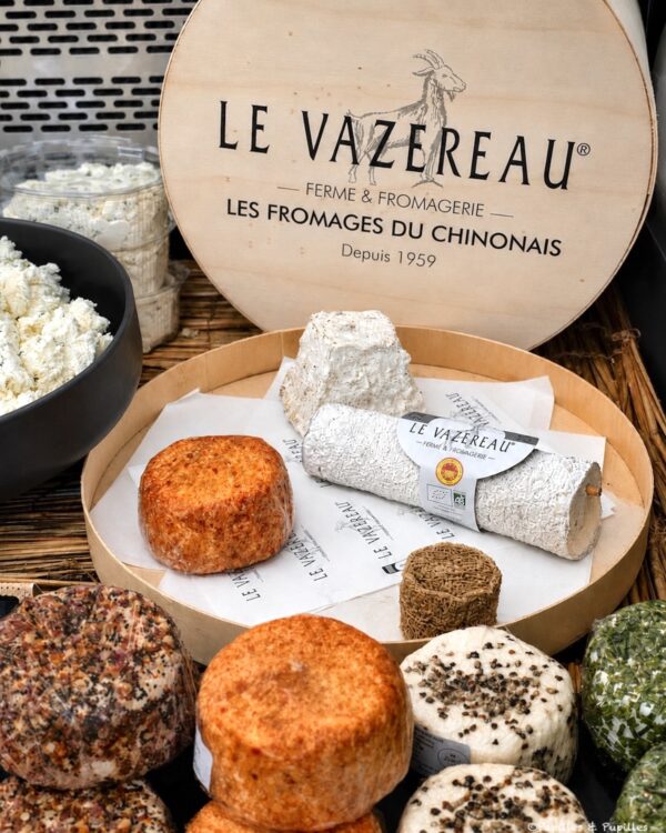Assorted cheeses on a round wooden board, including a wrapped log and orange-coated rounds, with a Le Vazereau sign in the background.