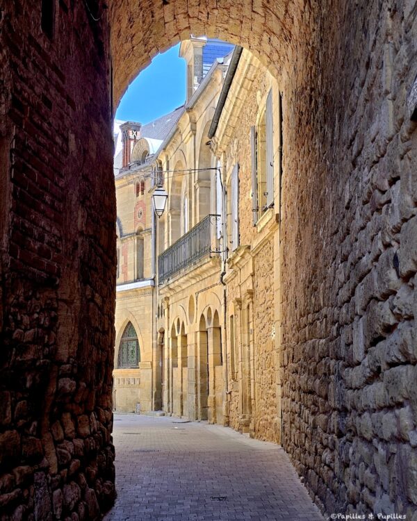 Narrow stone alley framed by tall historic buildings, with an arch overhead and blue sky beyond.