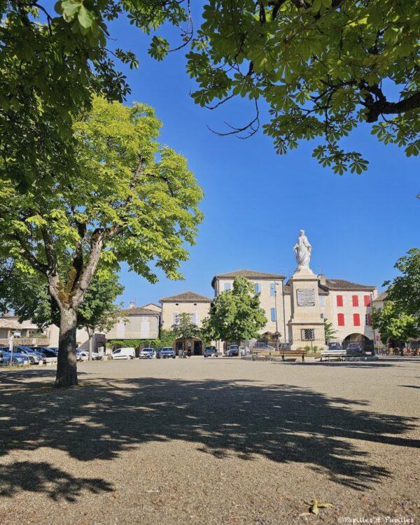 Place arborée de Castelnau-Montratier avec statue de la Vierge et maisons à arcades