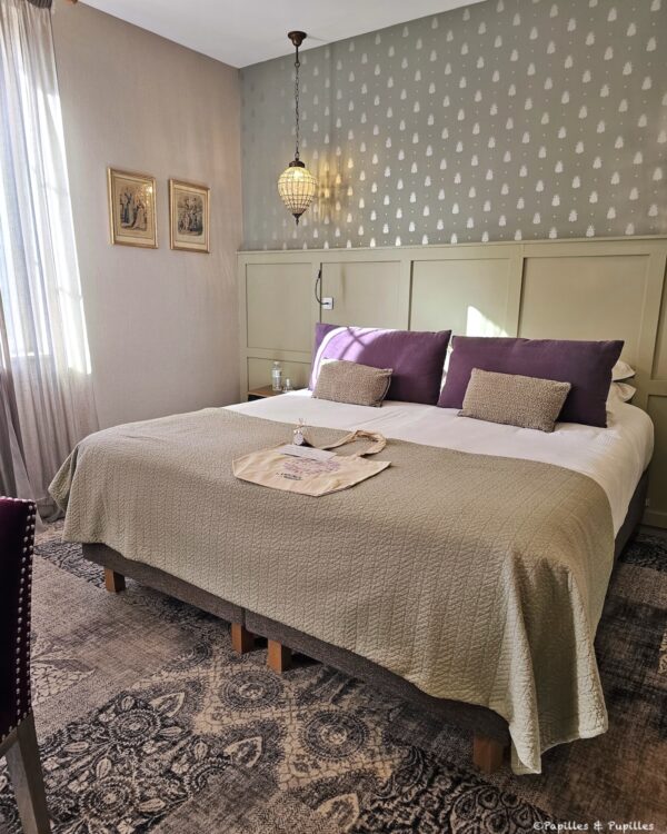 Lit double dans une chambre de l’hôtel Les Glycines avec coussins prune et papier peint à motifs