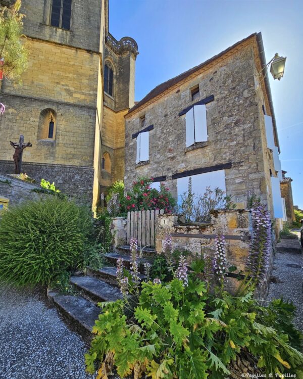 Maison en pierre fleurie au pied de l’église de Biron, avec volets blancs et végétation