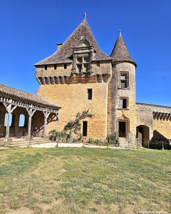 Façade du Château de Biron avec tour ronde, fenêtres sculptées et pierres blondes sous le ciel bleu