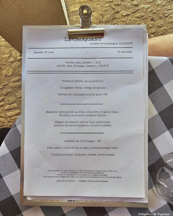 Menu on a clipboard outdoors, listing French dishes and prices on a white sheet against a stone wall and checkered tablecloth.