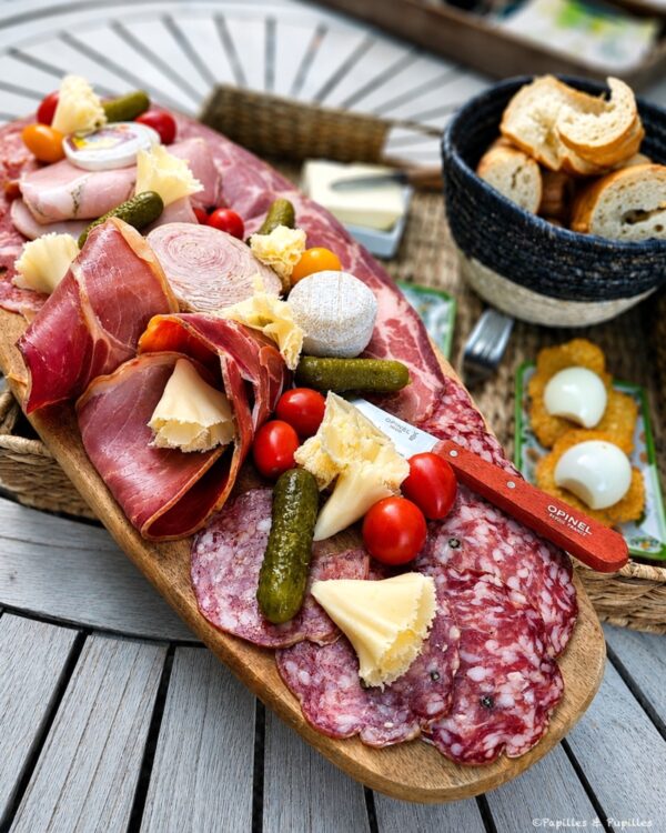 Charcuterie board with sliced cured meats, cheeses, cornichons, cherry tomatoes, and a basket of bread slices on a wooden surface.