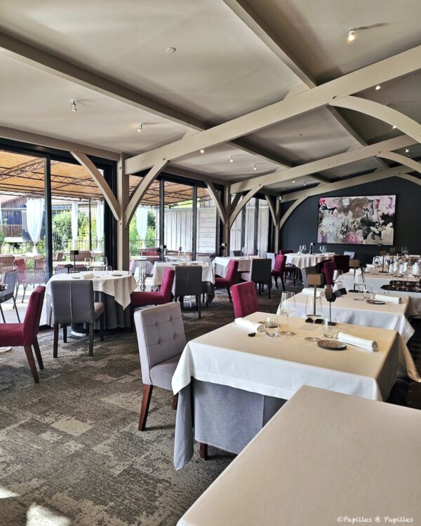 Elegant restaurant dining room with white-clothed tables, pink and gray chairs, large windows, and abstract painting on a dark wall.