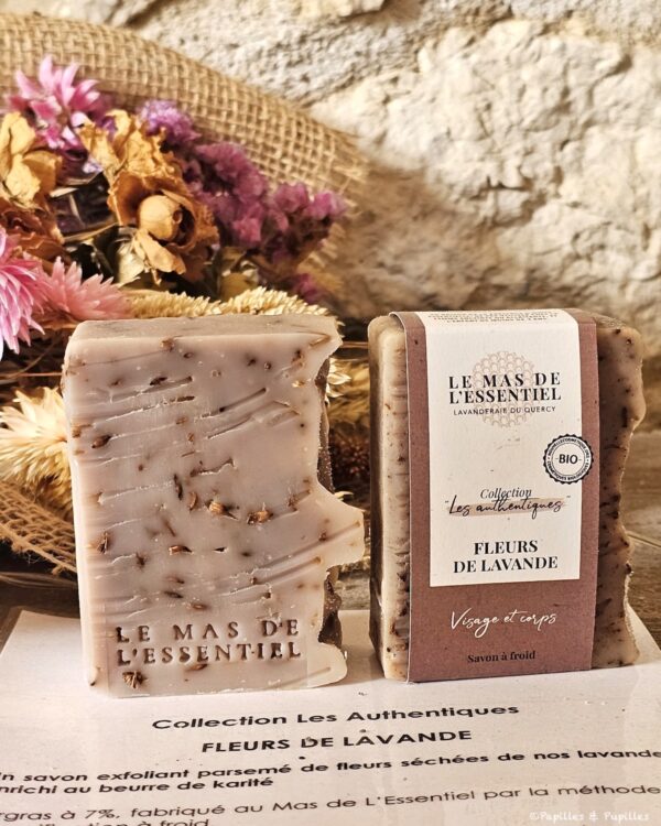 Two lavender-scented soaps on a surface, one unwrapped showing embedded plant bits and 'LE MAS DE L'ESSENTIEL' imprint, the other wrapped in paper with lavender label; dried bouquet in the background.