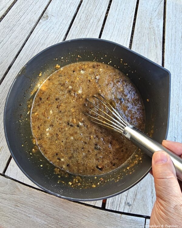 Whisk resting in a dark bowl with bubbling brown sauce on a wooden table, ready for stirring in.