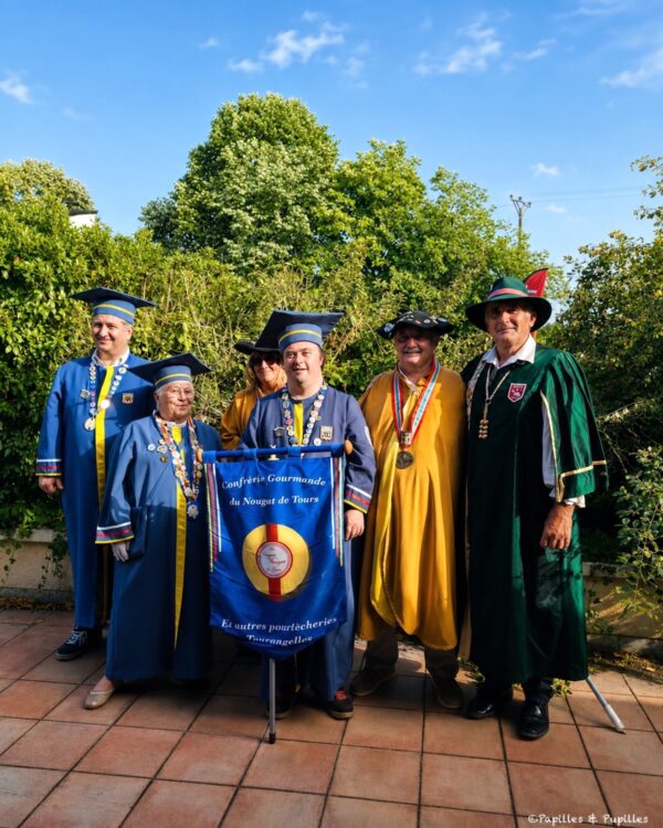 Group of people in ceremonial robes posing with a blue banner reading Confrérie Gourmande du Nougat de Tours outdoors.
