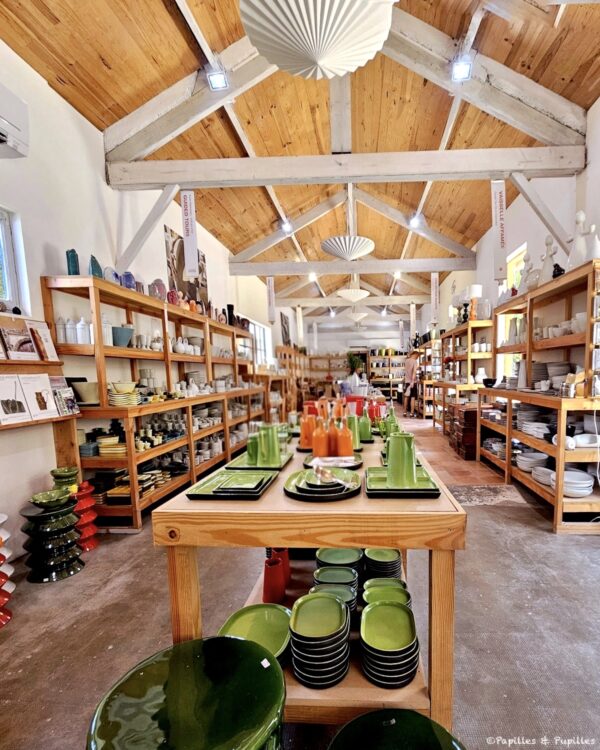 Bright pottery shop interior with wooden shelves full of ceramic dishes and glassware; a central table displays green dinnerware sets.